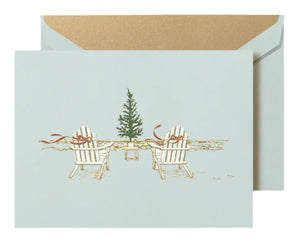 The Crane Seaside Christmas Card Set features two white Adirondack chairs with red ribbons by a festive tree on the shore; a gold-lined envelope completes this serene holiday scene.