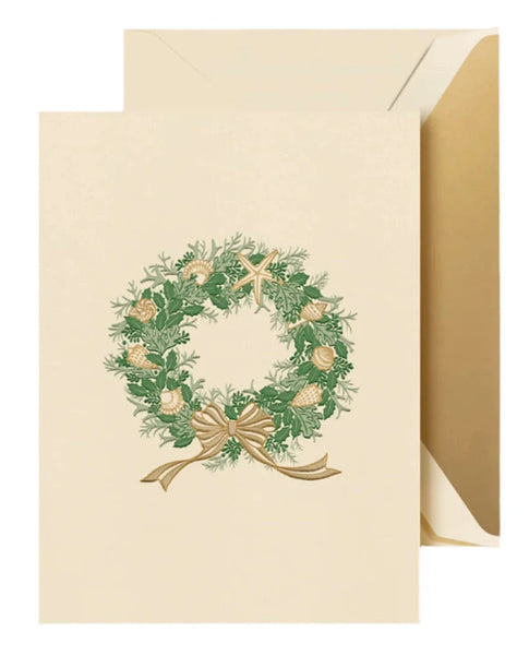 The Crane Seashells Wreath Card Set by Crane includes a beige holiday greeting card with an envelope, featuring a wreath design adorned with greenery, pine cones, and a gold bow. The card exudes a coastal tone reminiscent of sand dollars and seaside charm.