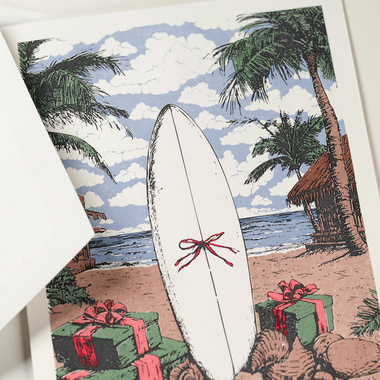 The Engraved Coastal Holiday Card Set by Crane features a surfboard with a red bow, gift boxes, palm trees, and a hut on the beach—ideal for sending seaside holiday cheer.