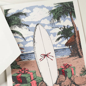 The Engraved Coastal Holiday Card Set by Crane features a surfboard with a red bow, gift boxes, palm trees, and a hut on the beach—ideal for sending seaside holiday cheer.
