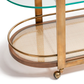 The Zodax Sloane Bar Cart features a glass top shelf, brass frame, woven rattan lower shelf, and small brass caster wheels—ideal for showcasing cocktail accessories in your home bar.