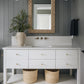 Modern bathroom featuring a white vanity, gold hardware, wall-mounted faucet, potted plant, two woven baskets, dark vertical wall paneling, and the Made Goods Gene Basket Weave Abaca Mirror adding subtle texture above the sink.