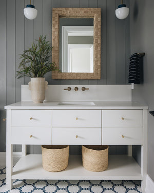 Modern bathroom featuring a white vanity, gold hardware, wall-mounted faucet, potted plant, two woven baskets, dark vertical wall paneling, and the Made Goods Gene Basket Weave Abaca Mirror adding subtle texture above the sink.