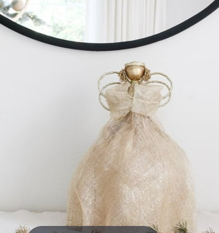 The Dekorasyon Sinamay Twine Angel Tree Topper, featuring gold wire and mesh, is elegantly displayed on a white surface before a round mirror and cream curtains—an ideal Christmas decoration.