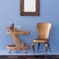 The Made Goods Lulu Retro Rattan Bar Cart with glass shelves stands beside a metallic gold chair, set against a light blue wall beneath a square decorative mirror.