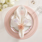 The Kim Seybert Printemps Napkin Set is displayed with a pink beaded ring on a pink and gold plate, beside a glass and white flowers, for an elegant spring table setting.