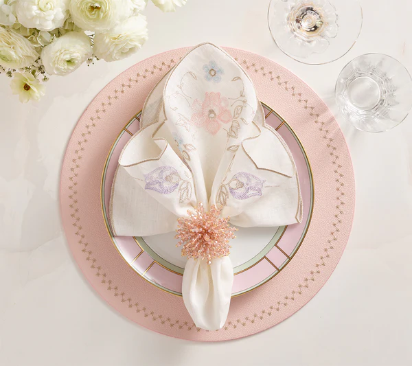 The Kim Seybert Printemps Napkin Set is displayed with a pink beaded ring on a pink and gold plate, beside a glass and white flowers, for an elegant spring table setting.