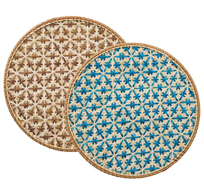 The Lanai Placemat Collection by Kim Seybert features two round rattan placemats with a star pattern—one in brown, one in blue and beige—both finished with natural fiber borders for added style.