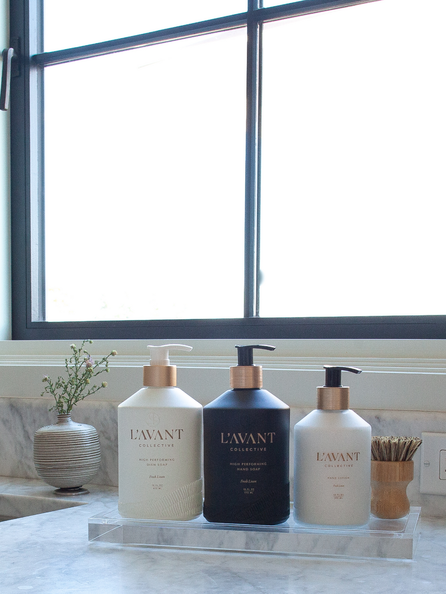 Three soap and lotion pump bottles on a L’Avant Collective Lucite Soap & Candle Tray resting on a marble countertop.