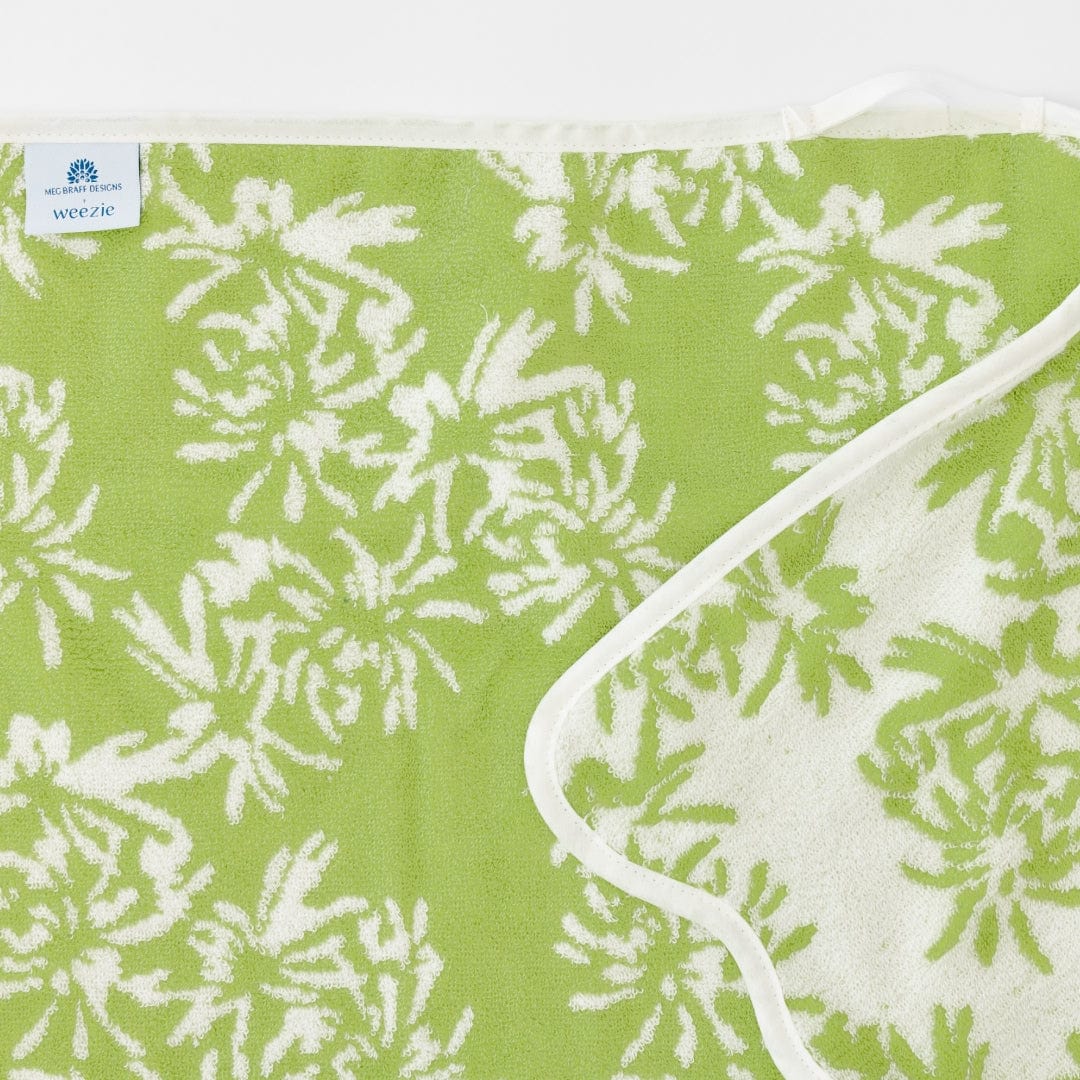 A beautifully folded green towel features a white floral pattern and scallop trim, with a small "Weezie" tag in the corner. This elegant piece is part of the exclusive Weezie Towels x Meg Braff Bath Collection, adding Palm Beach charm to your space.
