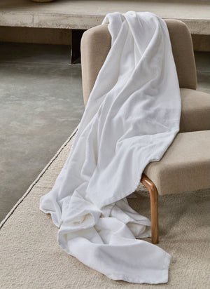 A Bovi White Finnley Blanket, made from 100% cotton, is draped over a beige upholstered chair and onto a light rug on a concrete floor—adding an inviting hint of premium European style.