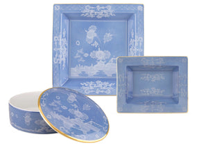 Three blue porcelain pieces from the Ginori 1735 Oriente Italiano Pervinca Collection—two square plates and a round lidded container with white floral designs and gold trim—are displayed against a white background.