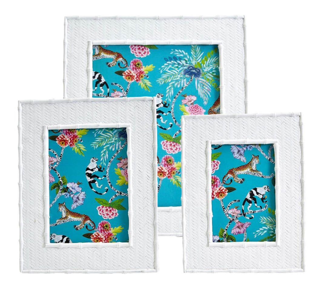 The Liana Frame Collection by Two's Company features three white textured photo frames with faux rattan and bamboo details, displaying vibrant animal prints, tropical flowers, and leaves on a bright blue background.