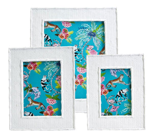 The Liana Frame Collection by Two's Company features three white textured photo frames with faux rattan and bamboo details, displaying vibrant animal prints, tropical flowers, and leaves on a bright blue background.
