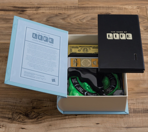 A box containing "The Game of Life Vintage Bookshelf Edition" by WS Game Company. The open box reveals the game board, instructions, and carefully arranged game pieces inside. Featuring vintage graphics, the box rests on a wooden surface.