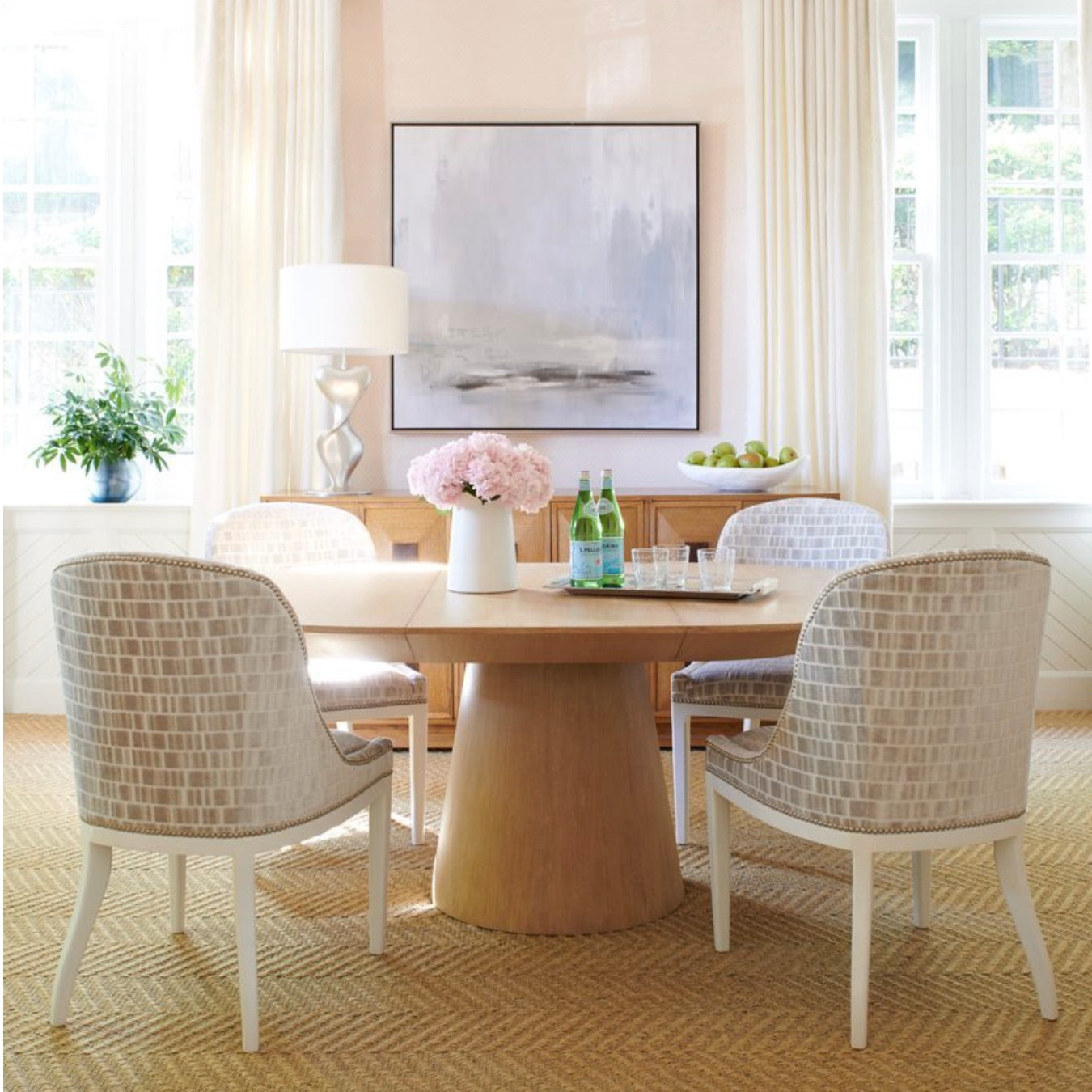 The Sherrill/Mr & Mrs Howard Discus Extendable Dining Table with four patterned chairs, a vase of pink flowers, water bottles, and glasses sits in a bright, modern room with large windows, artwork, and a sideboard.