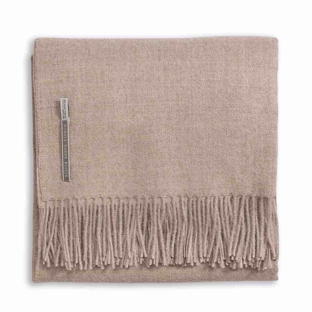 Alicia Adams Alpaca's Classic Throw, a folded beige blanket from the Alicia Adams Alpaca collection, is crafted from cashmere and baby alpaca, featuring fringes on the bottom edge and a small fabric tag on the left side. It is fair-trade made in Peru.