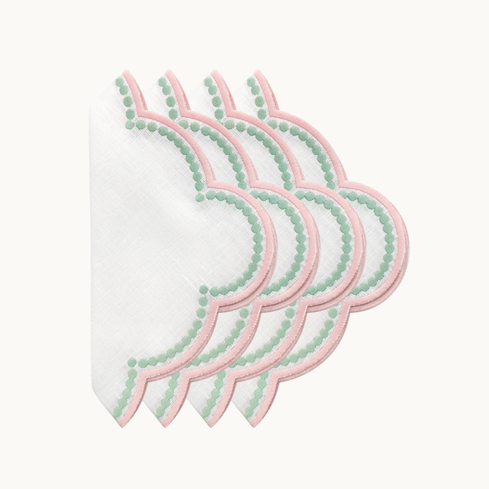 Four white linen napkins from the Duggan Society’s Lilibeth Table Linen Collection, with scalloped edges, pink borders, and green dot accents, are fanned out on a plain background for an elegant decorative touch.