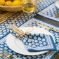 A white plate with a blue-patterned Pomegranate Inc Pom Buti Denim Collection napkin tied with straw, flanked by gold cutlery, offers a charming dining experience. A wine glass and a bowl of lemons in the background add to the inviting scene, enhanced by the subtle elegance of floral design accents.
