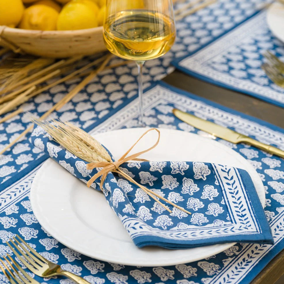 A white plate with a blue-patterned Pomegranate Inc Pom Buti Denim Collection napkin tied with straw, flanked by gold cutlery, offers a charming dining experience. A wine glass and a bowl of lemons in the background add to the inviting scene, enhanced by the subtle elegance of floral design accents.