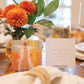 A table with Anne Neilson: Love Scripture Cards by Anne Neilson, two display cards, orange flowers in a vase, and table settings. One card shows inspirational art, the other displays a Bible verse from Colossians 3:14.