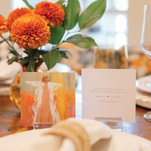 A table with Anne Neilson: Love Scripture Cards by Anne Neilson, two display cards, orange flowers in a vase, and table settings. One card shows inspirational art, the other displays a Bible verse from Colossians 3:14.