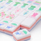 The Oh My Mahjong Mahjong Tile Collection features stylish pastel tiles with floral, animal, and plant designs, neatly arranged with one hand-painted tile displayed in the foreground.
