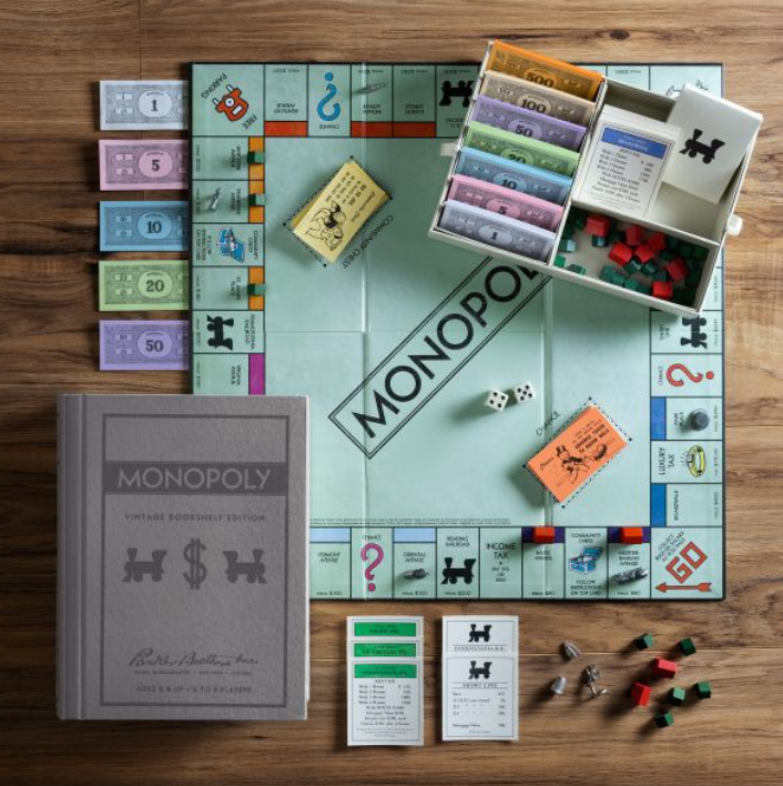 A Monopoly Vintage Bookshelf Edition setup with various game cards, money, houses, hotels, dice, and metal tokens is laid out on a wooden surface. The edition from WS Game Company includes the game box and a gray book-style case.