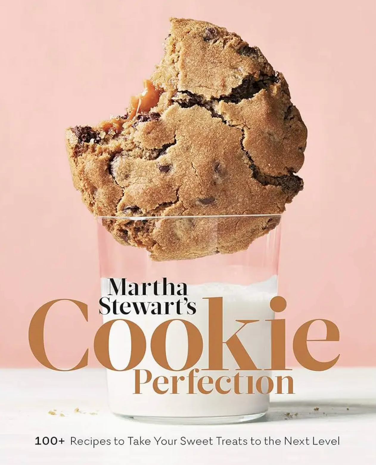 A large chocolate chip cookie is partially dipped in a glass of milk against a light pink background. The text features the product name, “Martha Stewart’s Cookie Perfection” by Penguin Random House, inviting you to explore creative cookies and delightful recipes for every occasion.