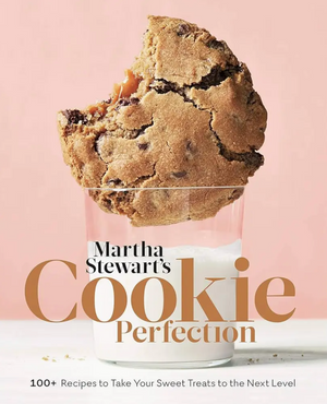 A large chocolate chip cookie is partially dipped in a glass of milk against a light pink background. The text features the product name, “Martha Stewart’s Cookie Perfection” by Penguin Random House, inviting you to explore creative cookies and delightful recipes for every occasion.