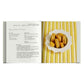 An open Simon & Schuster Ladurée Macarons cookbook displays a recipe on the left, while a plate of yellow Ladurée macarons sits on the right, all arranged on a yellow-striped surface.