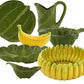 The Bananas From Madeira Serveware Collection by Bordallo Pinheiro features ceramic plates, bowls, a pitcher, and a banana-shaped dish, all inspired by the banana tree with green leaf and yellow banana designs.