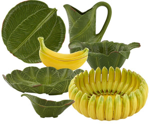 The Bananas From Madeira Serveware Collection by Bordallo Pinheiro features ceramic plates, bowls, a pitcher, and a banana-shaped dish, all inspired by the banana tree with green leaf and yellow banana designs.