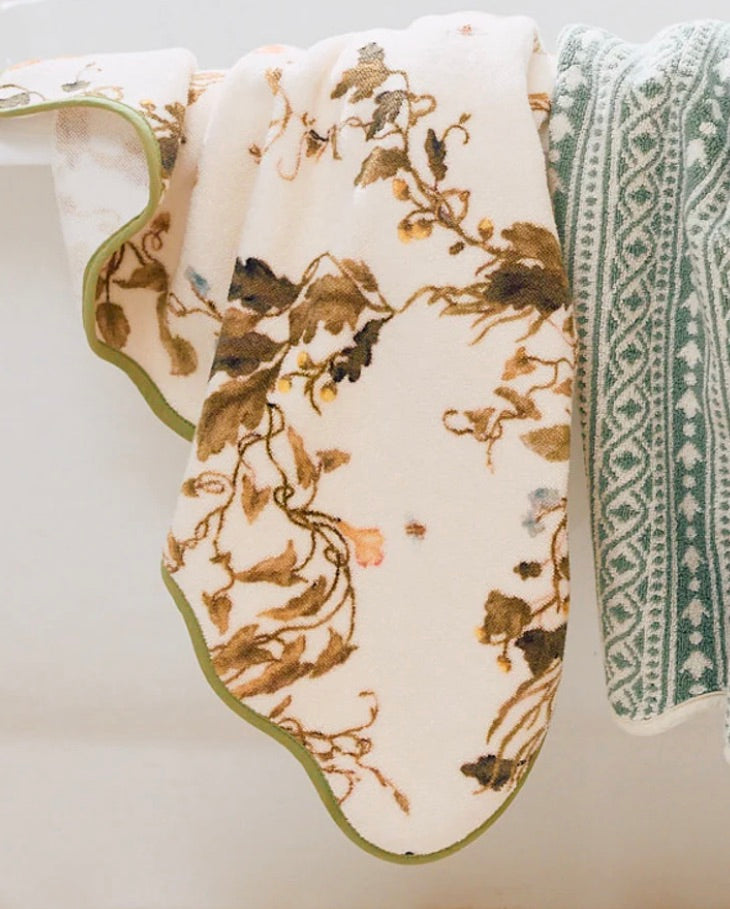 Two towels hang side by side: a luxury bath towel with a green and white geometric pattern, and the Vivian Magnolia Floral Scallop Bath Towel by Weezie Towels, featuring a brown and green floral design on white.