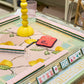 The Oh My Mahjong Tile Collection delivers a vibrant Mahjong gaming scene on a lemon-patterned table, featuring hand-painted tiles, racks, a yellow lamp, pen, red pouch, and glass.