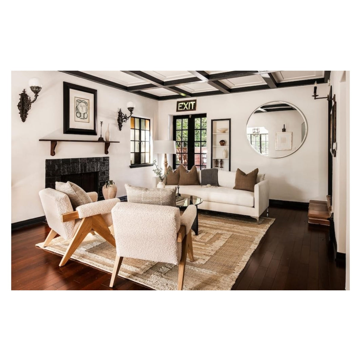 A modern living room from Meridith Baer: The Making of a Home by Abrams features a white sofa, armchairs, glass coffee table, wood floors, and a bold “EXIT” sign above the window.