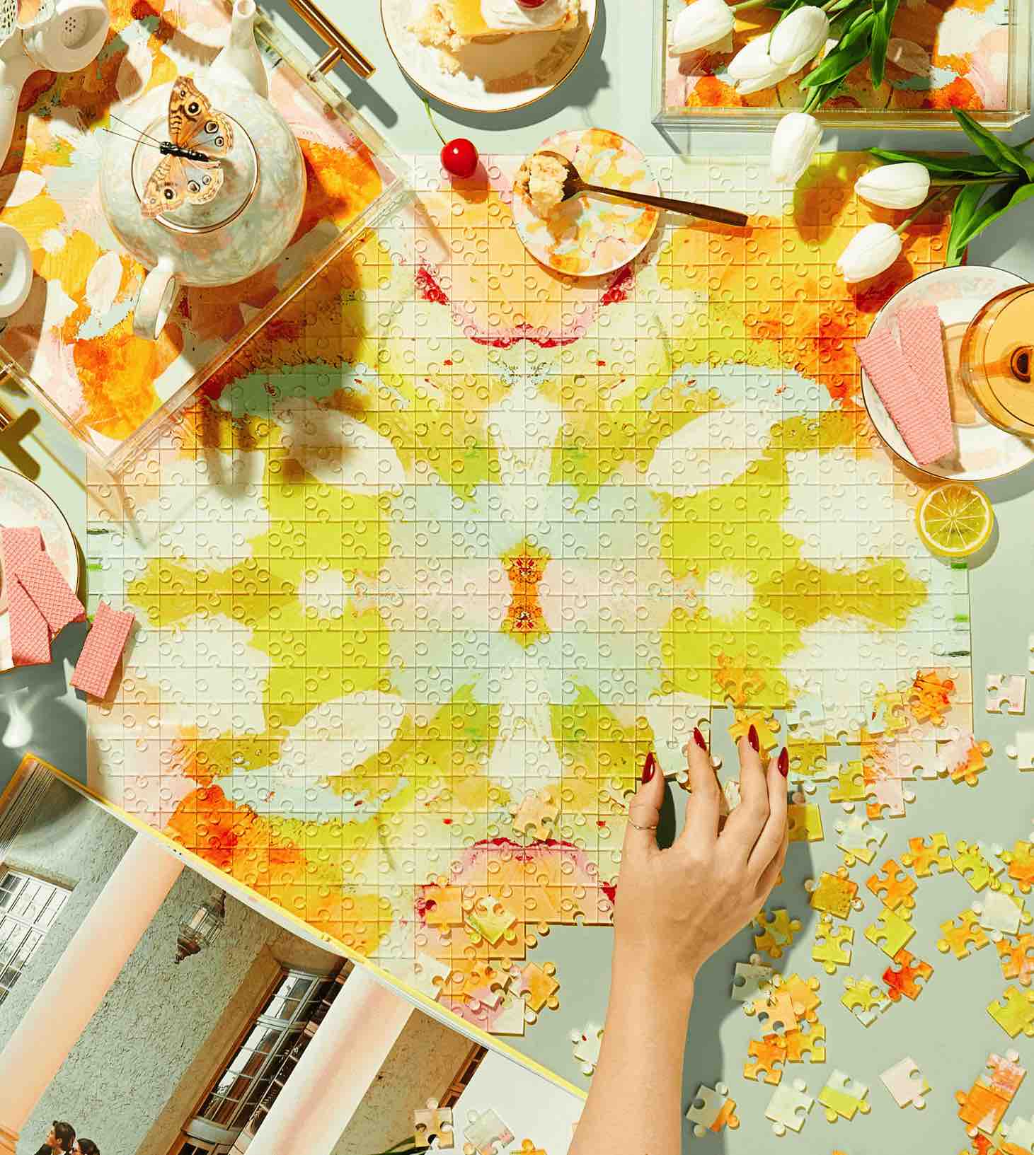 A hand arranges pieces of the colorful Acrylic Puzzle by Tart by Taylor on a table with tulips, desserts, drinks, and scattered puzzle pieces—an eye-catching touch for vibrant home decor.