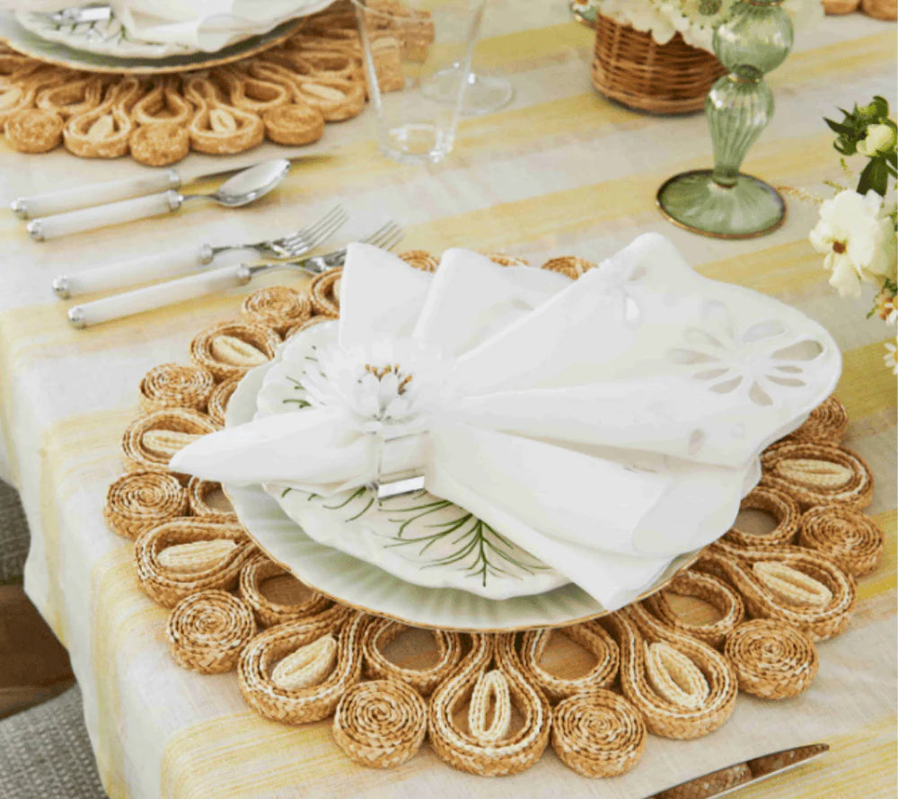 A table setting features a woven placemat, white plates, the Kim Seybert Bloom White Napkin Set with a floral napkin ring, and silver utensils atop a striped tablecloth.