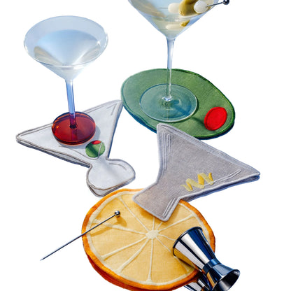Two martini glasses rest on Piecework Martini Cocktail Napkins shaped like martini glasses, set atop circular coasters resembling an olive and a citrus slice, alongside a cocktail pick and jigger.