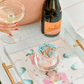 A hand holding champagne next to a Veuve Clicquot bottle, the Acrylic Match Striker Collection by Tart by Taylor with blue-tipped matches, and a floral tray come together under a white umbrella for the ideal coffee table setup.