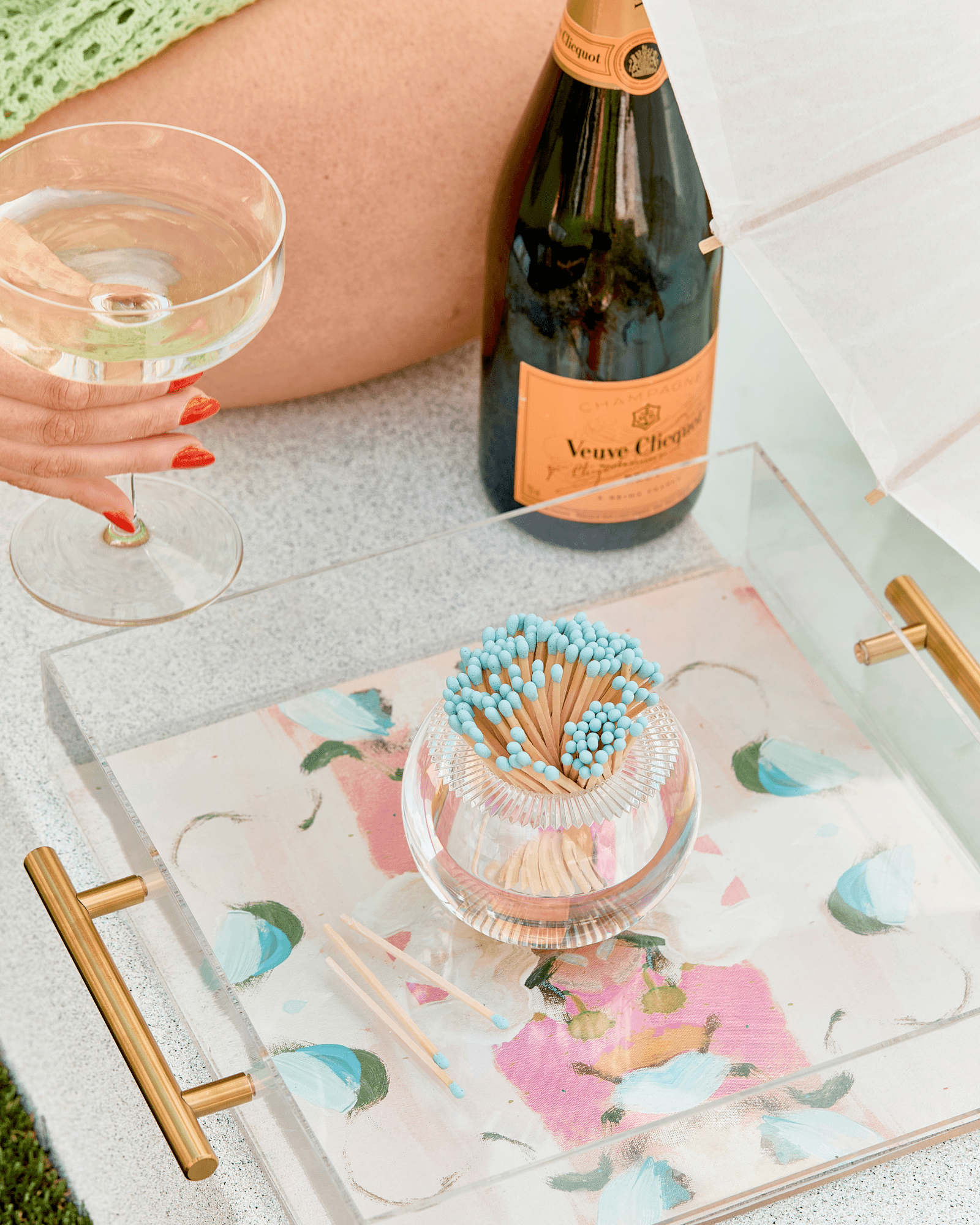 A hand holding champagne next to a Veuve Clicquot bottle, the Acrylic Match Striker Collection by Tart by Taylor with blue-tipped matches, and a floral tray come together under a white umbrella for the ideal coffee table setup.