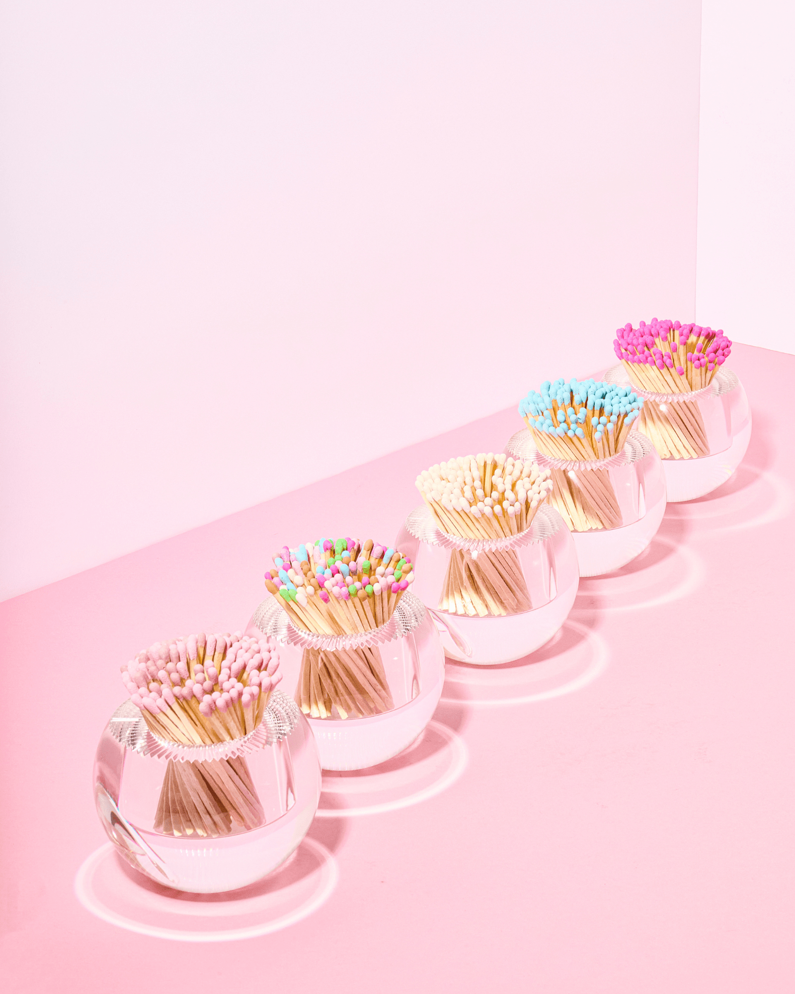 The Acrylic Match Striker Collection by Tart by Taylor features five round glass bowls with colorful-tipped wooden matches, lined up on a pink surface—ideal as a playful coffee table accessory and perfect for use with any acrylic match striker or strike pad.