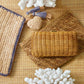 An Aerin Allegra Raffia Pouch in a neutral woven chevron stitch rests on two flat woven pouches, accented by a small heart-shaped charm and tassel on a textured surface.
