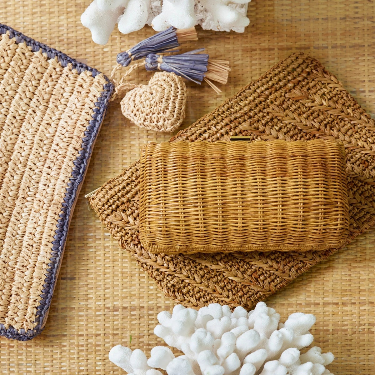 An Aerin Allegra Raffia Pouch in a neutral woven chevron stitch rests on two flat woven pouches, accented by a small heart-shaped charm and tassel on a textured surface.