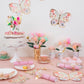 A pastel bridal shower table with butterfly decor, a cake, Meri Meri Pink Tissue Floral Crackers, floral cups, pastries, and gold cutlery arranged on a pink tablecloth.