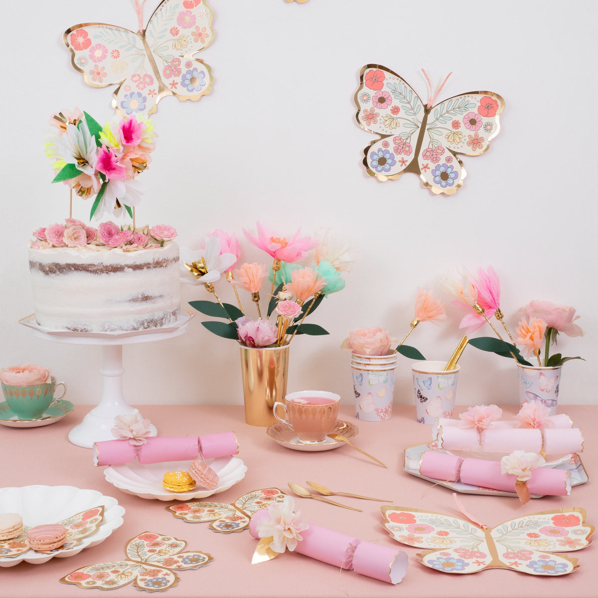 A pastel bridal shower table with butterfly decor, a cake, Meri Meri Pink Tissue Floral Crackers, floral cups, pastries, and gold cutlery arranged on a pink tablecloth.
