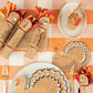 Thanksgiving table setting featuring Meri Meri Turkey Party Crackers, turkey-themed napkins, festive party favors, paper turkey decorations, and utensils arranged on an orange and white checkered tablecloth.