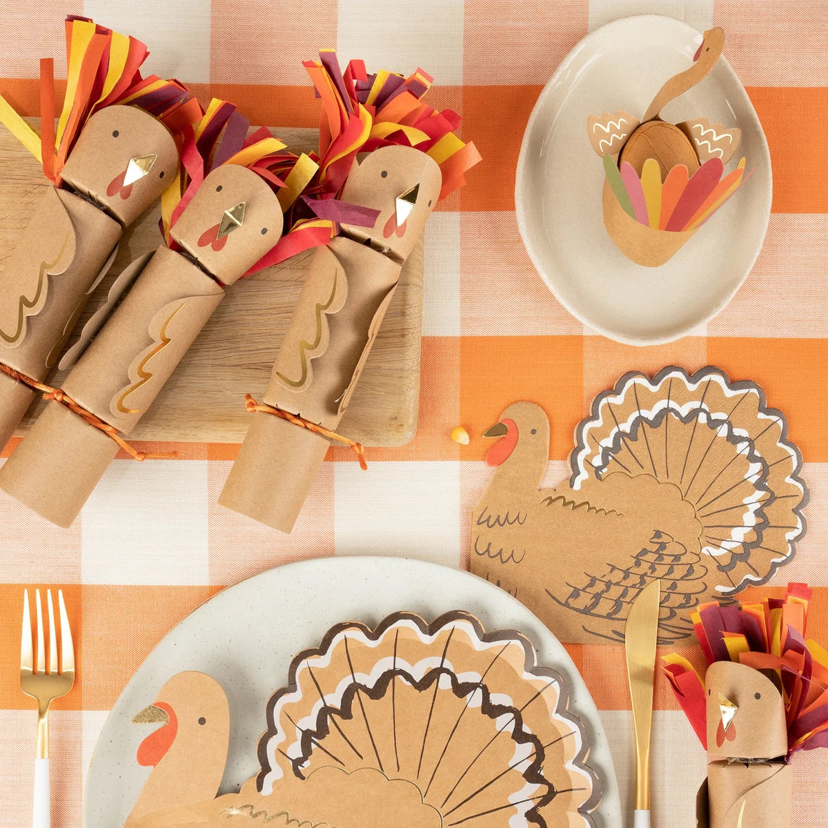 Thanksgiving table setting featuring Meri Meri Turkey Party Crackers, turkey-themed napkins, festive party favors, paper turkey decorations, and utensils arranged on an orange and white checkered tablecloth.
