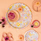 Scalloped floral-patterned plates and bowls, each with pink and peach flowers, are arranged on a peach-toned surface next to the Rainbow Garden Tray by Meri Meri for a charming decorative touch.