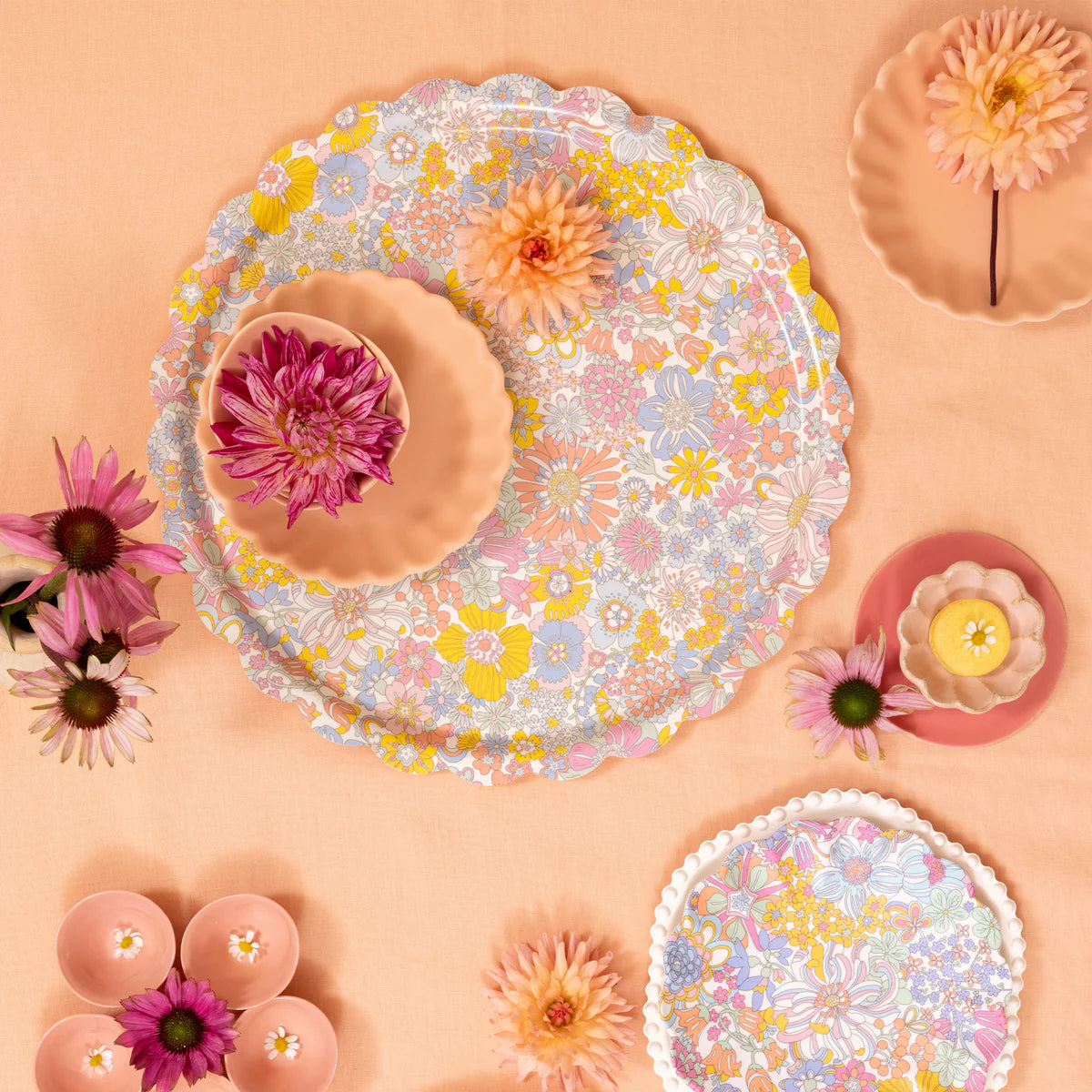 Scalloped floral-patterned plates and bowls, each with pink and peach flowers, are arranged on a peach-toned surface next to the Rainbow Garden Tray by Meri Meri for a charming decorative touch.